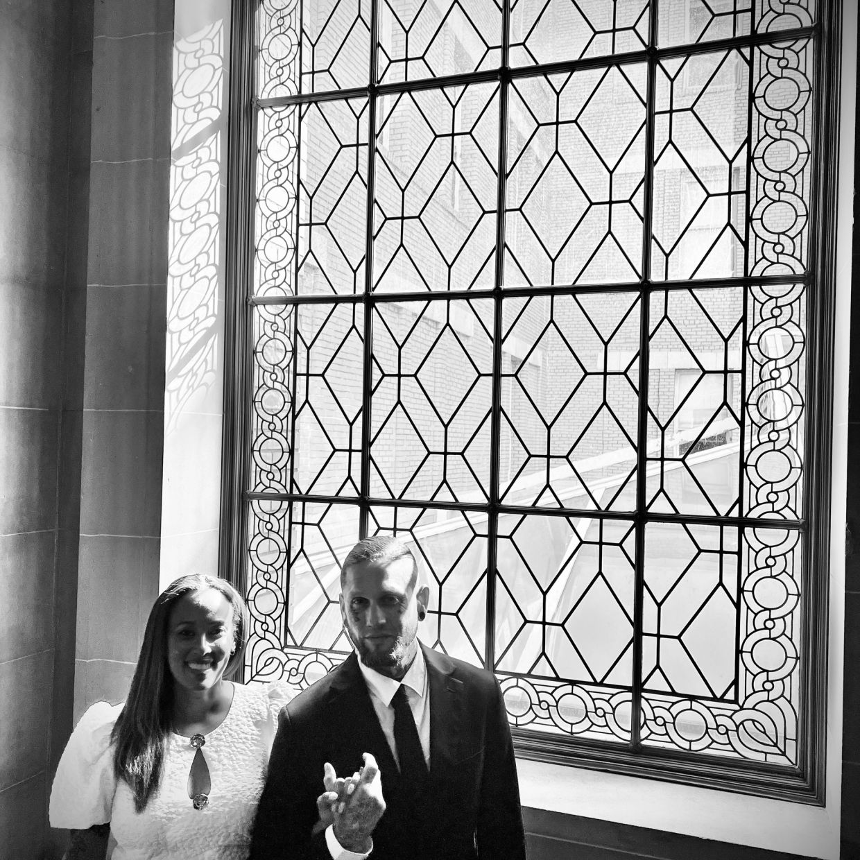 Couple posing by an ornate window, dressed formally.