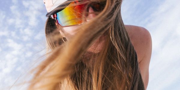 Person with long hair wearing reflective sunglasses and a cap against a cloudy sky.