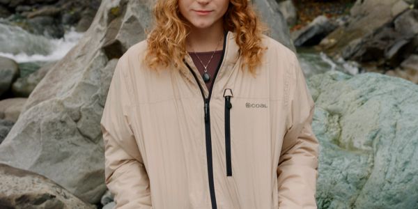 Person in a beige jacket standing by rocky river scenery.