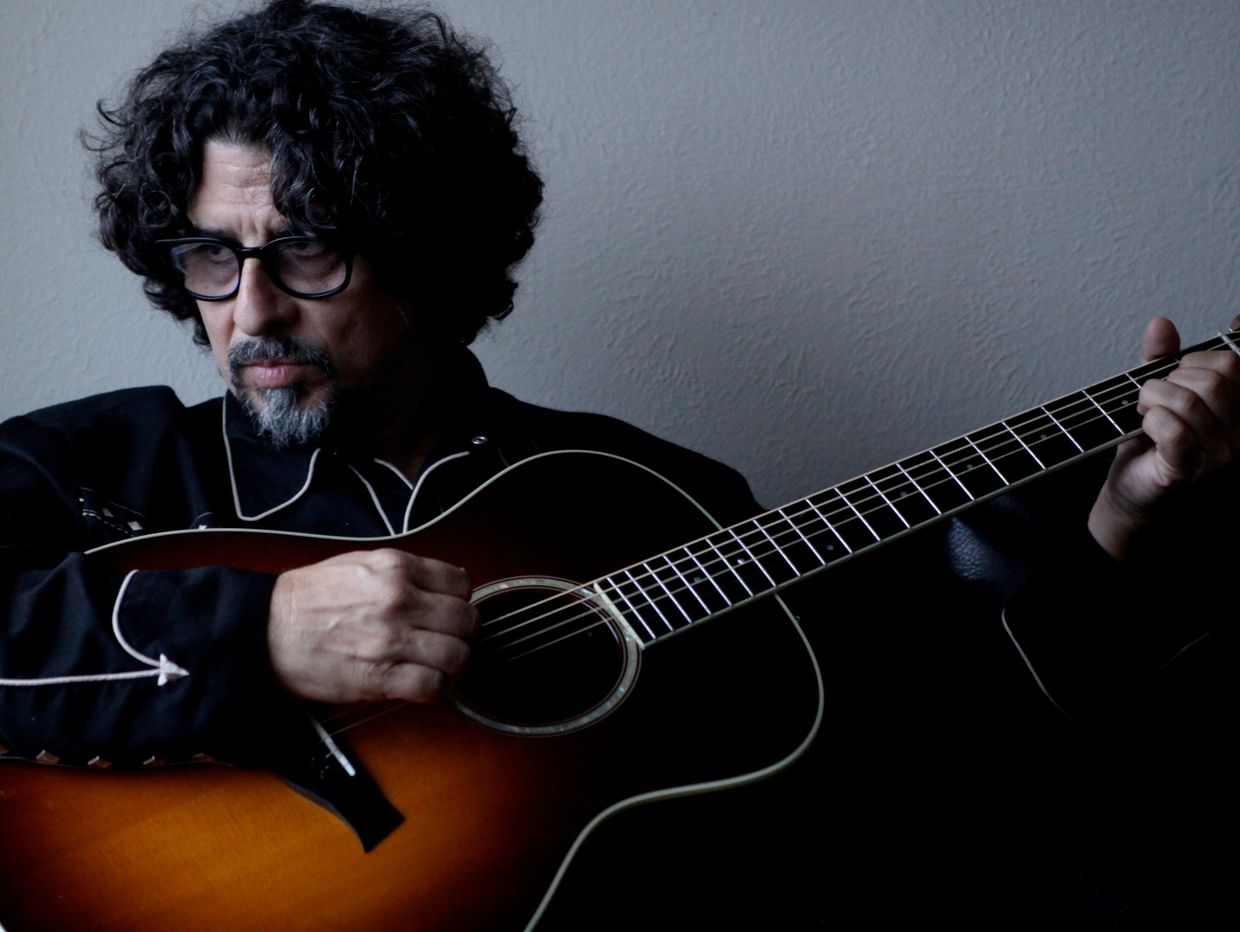 Man with curly hair playing an acoustic guitar in a dimly lit room.
