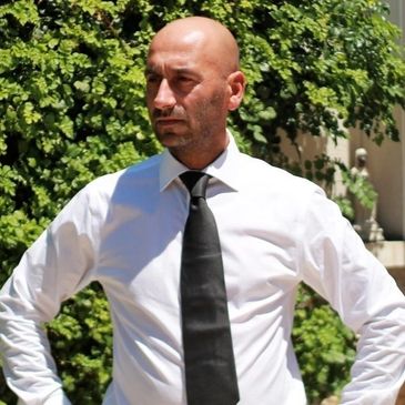 Man in white shirt and black tie standing outdoors with hands on hips.