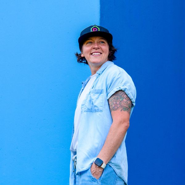 Person smiling against a two-tone blue wall, wearing a cap and showing a floral tattoo.