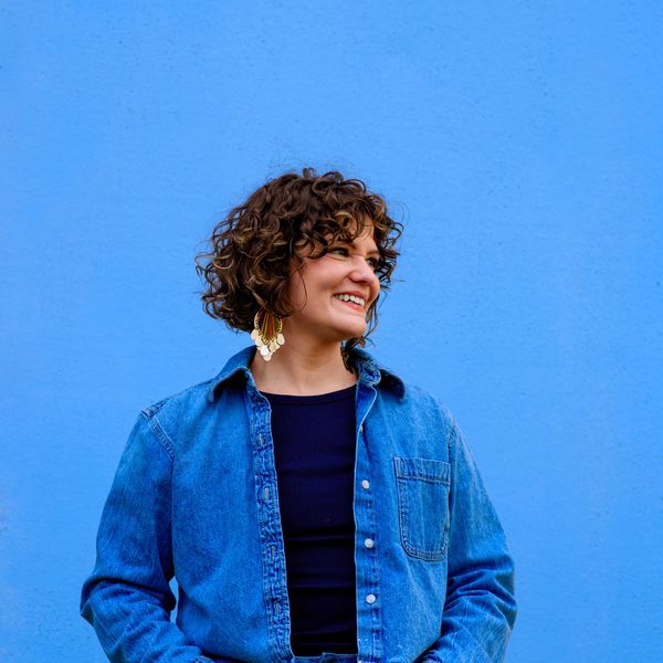 Smiling woman in denim jacket against a bright blue wall.