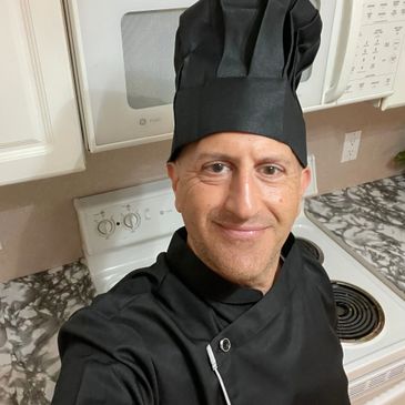 Smiling man wearing a black chef hat and coat in a kitchen.