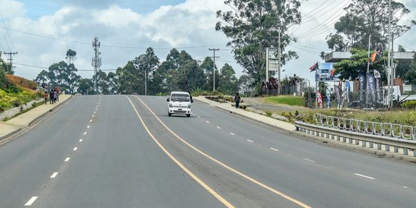 A white van drives on a wide, empty multi-lane road with few pedestrians nearby.