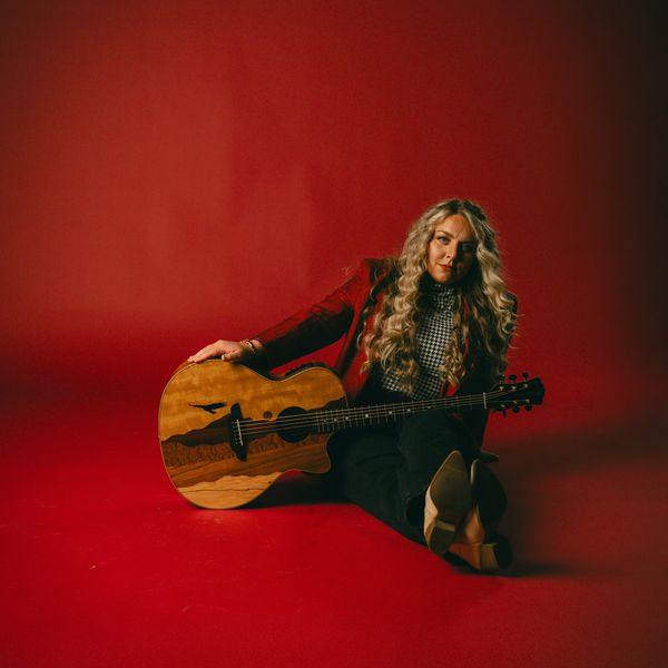 Woman sitting with guitar against red backdrop.