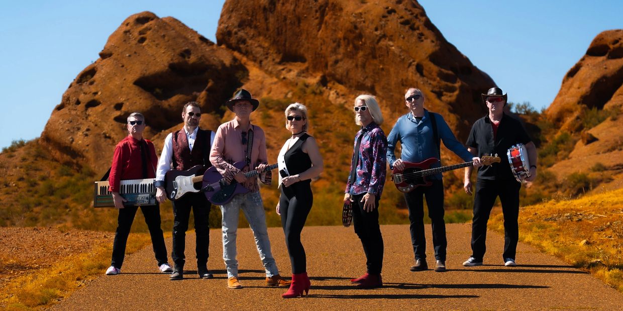 Band of seven musicians posing in a desert landscape with instruments.