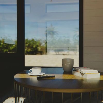 Coffee cup, mug, books, and remote on a round wooden table by a sunlit window.
