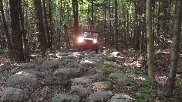 Off-road vehicle navigating rocky forest trail with headlights on.