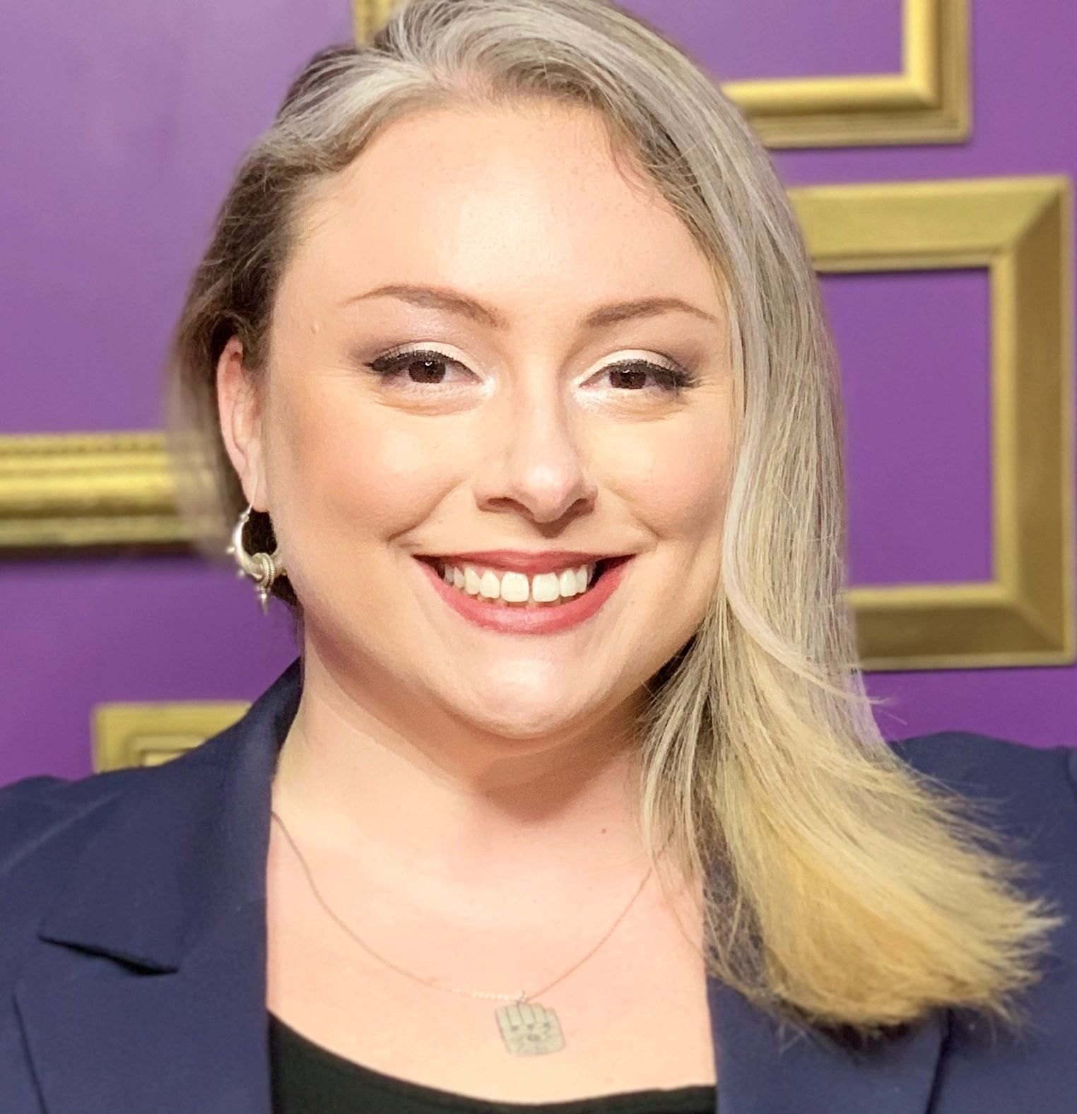 Smiling woman with blonde hair against a purple wall with gold frames.