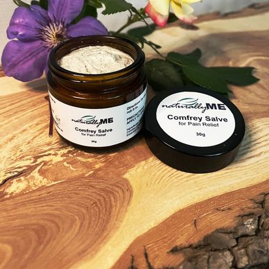 A jar of NaturallyME Comfrey Salve for pain relief with colorful flowers on a wooden surface.