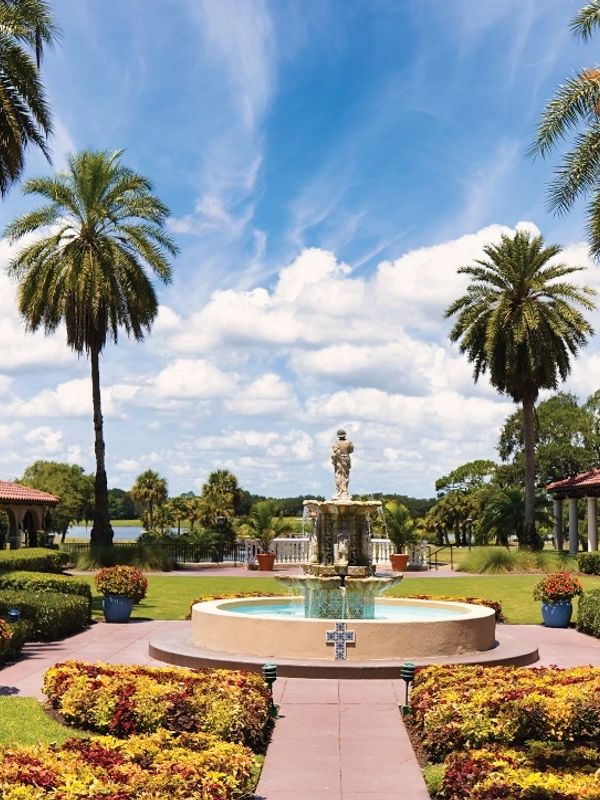 A garden with a central fountain, palm trees, and a bright blue sky.