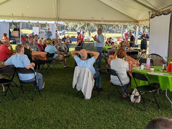 Community meeting held under a large outdoor tent with attendees seated and listening.