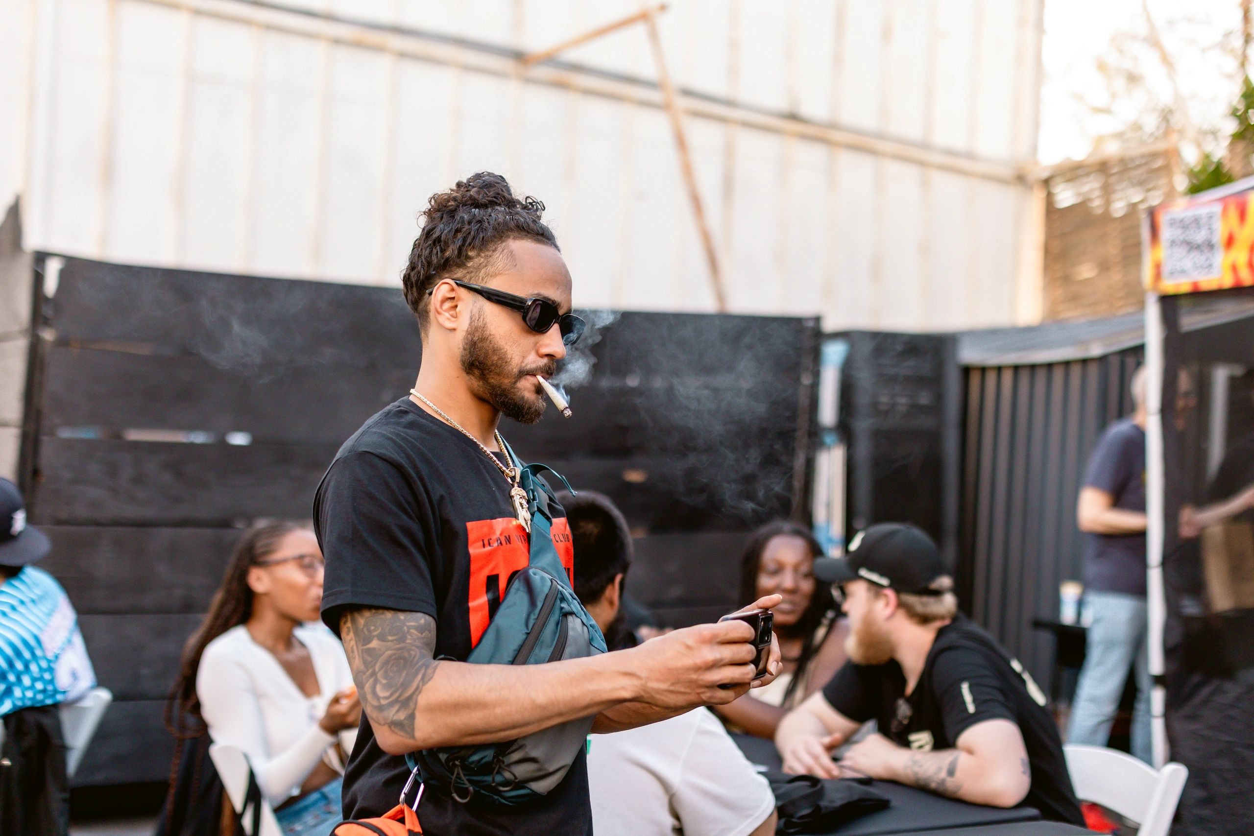 Man smoking and using phone outdoors with people in background.