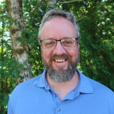 Smiling man with glasses and beard wearing a blue polo shirt outdoors.