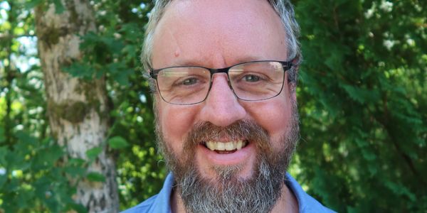 Smiling man with glasses and beard wearing a blue polo shirt outdoors.