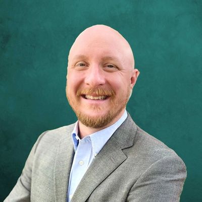 Smiling bald man with a beard in a gray suit jacket against a teal background.