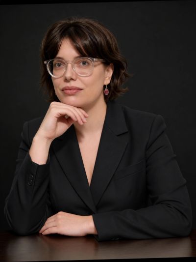 Confident woman with glasses in a black blazer posing against a dark background.