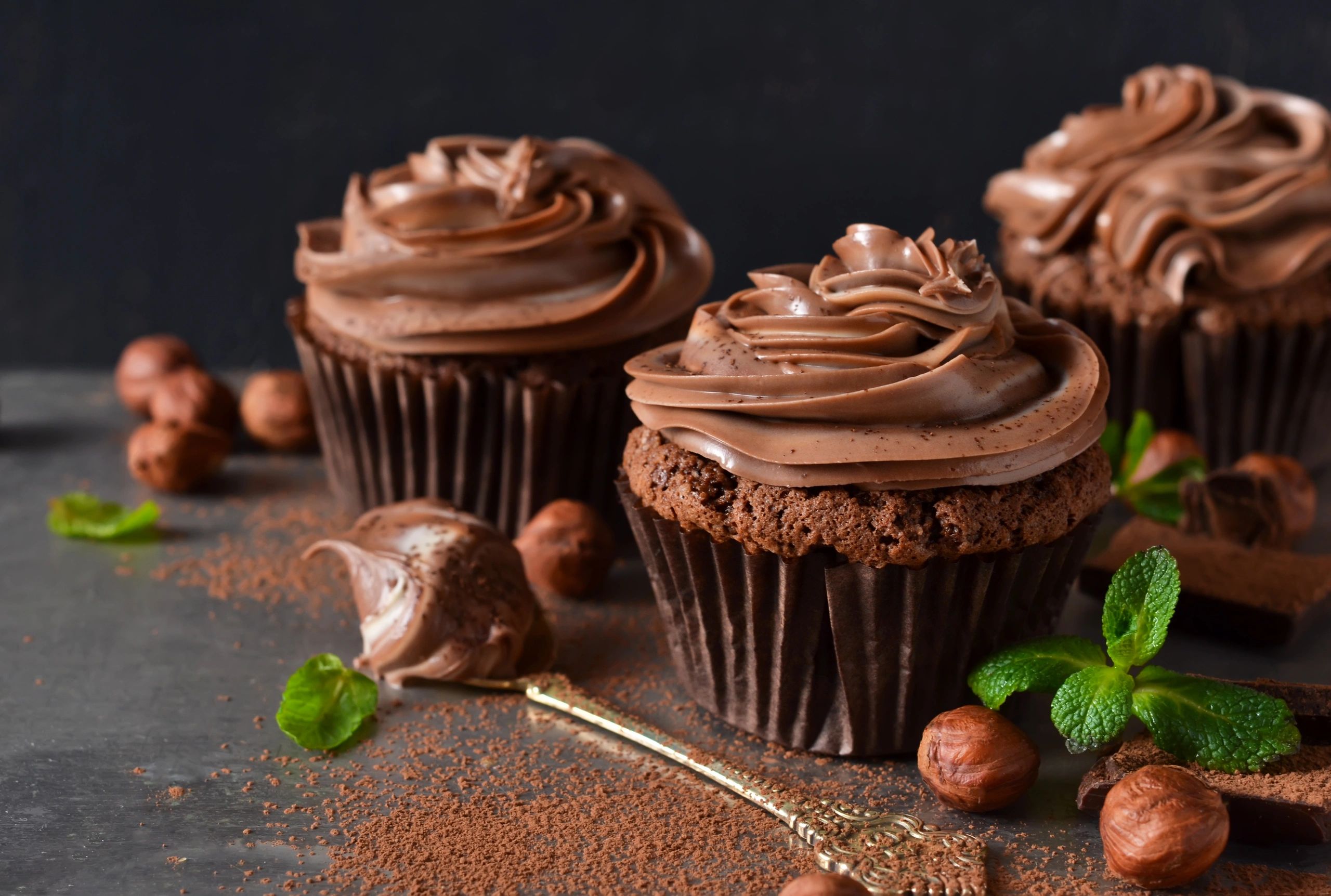 Chocolate cupcakes with creamy frosting and hazelnuts on a dark surface.