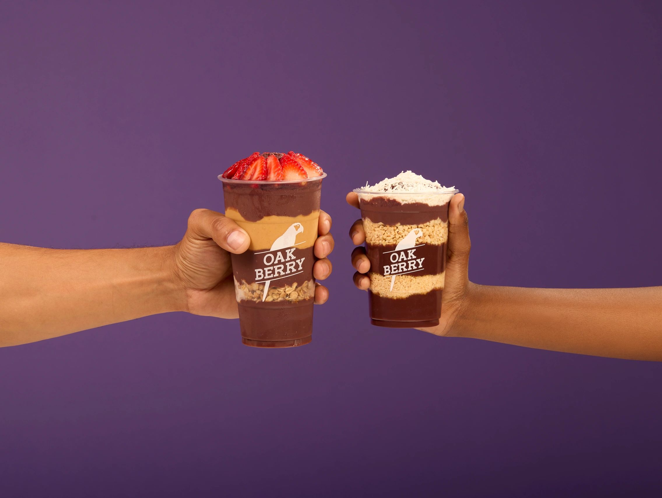 Two hands holding Oak Berry dessert cups with toppings against a purple background.