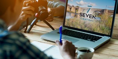 A laptop on a desk with seven central branding on it. two creative people are looking at the screen