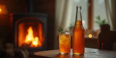 A bottle of chilled ginger beer on a rustic table in front of a wood burning stove in the winter