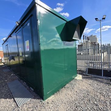 Green industrial electrical enclosure under a blue sky near a fenced power substation.
