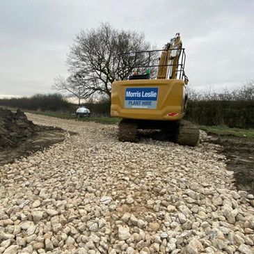 Excavator laying a gravel path in a rural area under a cloudy sky.