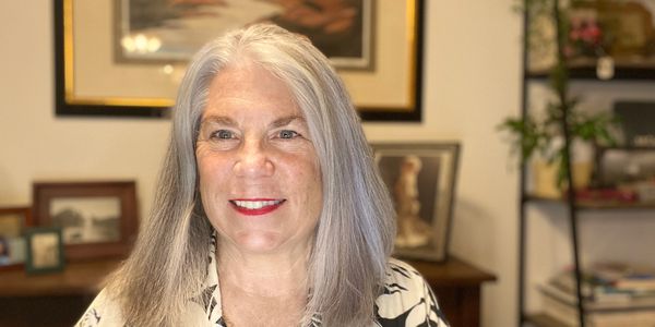 Platinum grey haired smiling woman on zoom call. There are framed photos and a bookshelf with plants