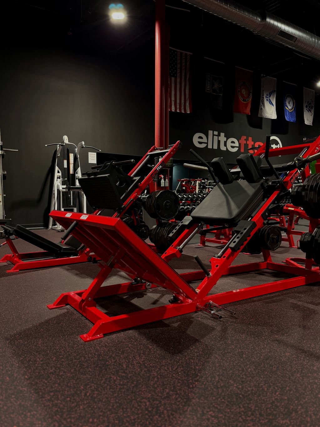 Red leg press machine in a dimly lit gym with flags and weights.