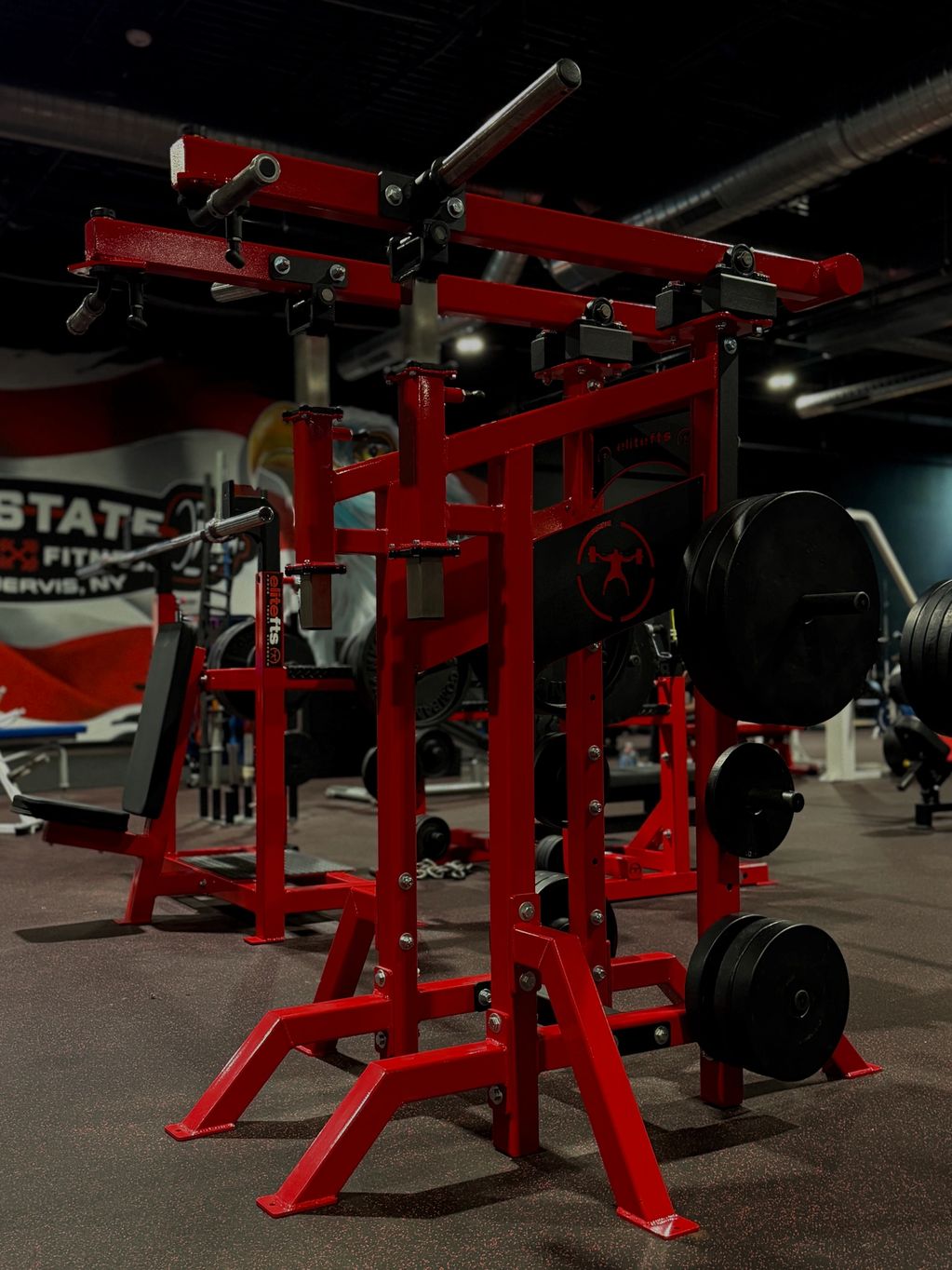 Red gym squat rack with weight plates in a fitness center.
