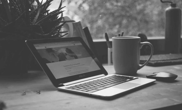 Black and white photo of a laptop, coffee mug, and mouse on a desk by a window.