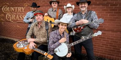 Six country musicians with instruments posing against a brick wall.