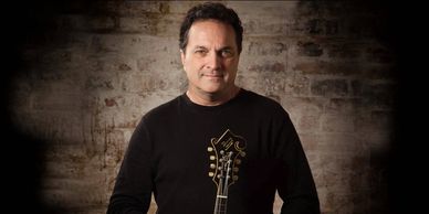 Man holding guitar in front of a textured brick wall.