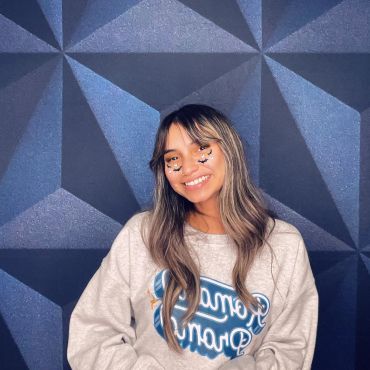 Smiling young woman with butterfly face stickers, wearing a cozy sweatshirt against a geometric wall.