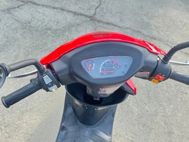 View of a red scooter's dashboard and handlebars on a paved surface.