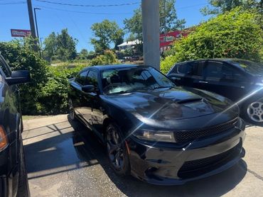 Black sports car parked outdoors with greenery and blue sky.