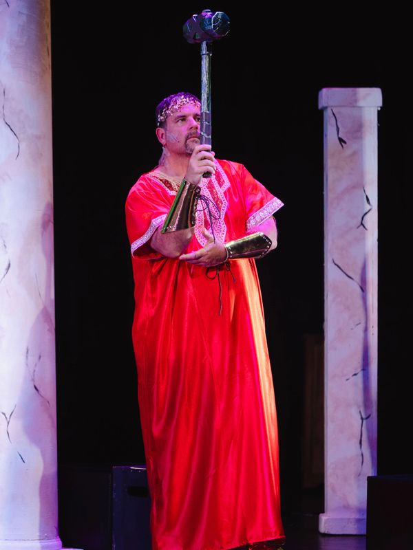 Man in red robe and golden crown holding a large hammer on stage.