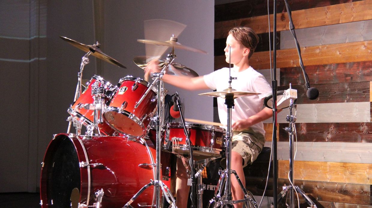 A young boy energetically playing a red drum set on stage.