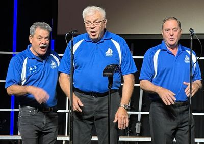 Three men in matching blue shirts singing on stage with microphones.