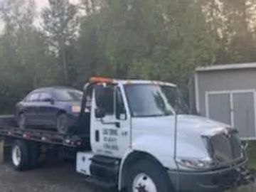 A flatbed tow truck carrying a dark-colored sedan in a wooded area.