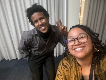 Two friends smiling, one making a peace sign gesture indoors.
