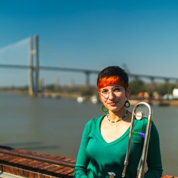 Person holding a trombone near a waterfront with a bridge in the background.