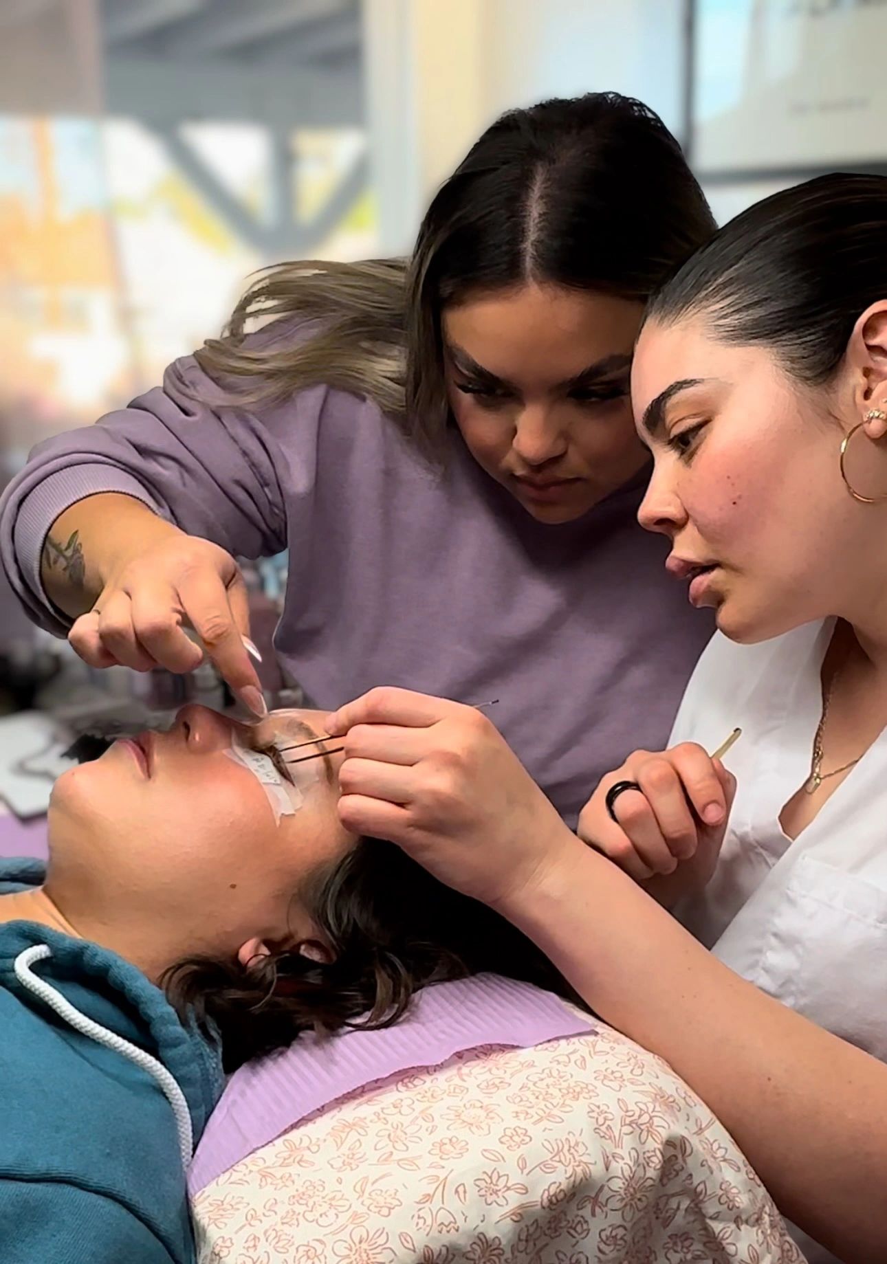 Two women carefully applying eyelash extensions on a lying client.