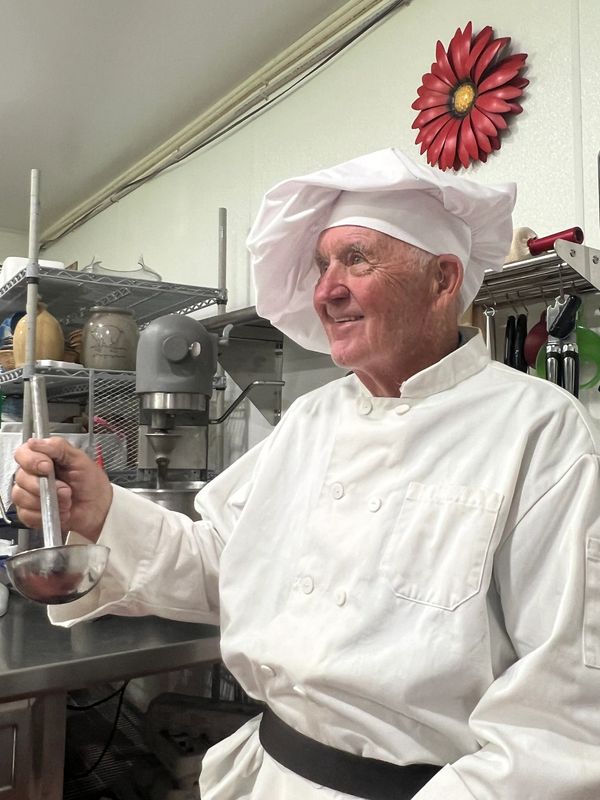 Elderly chef in white uniform holding a ladle in a kitchen.