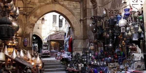 A vibrant market alley filled with traditional lamps and crafts under an ancient stone archway.