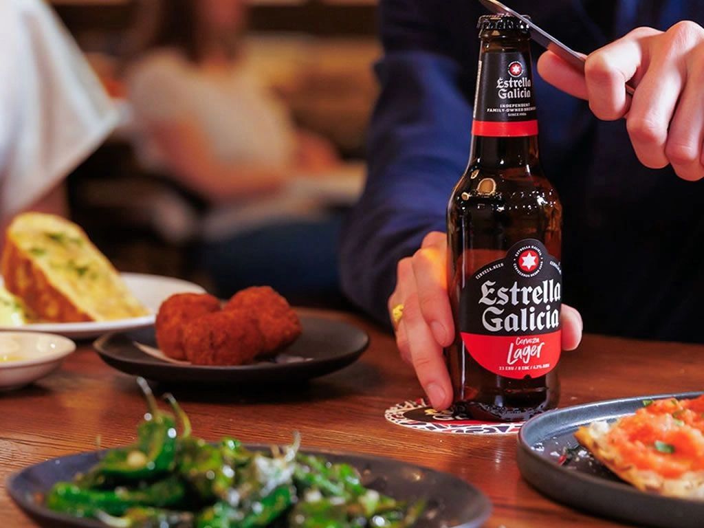 Person opening an Estrella Galicia beer bottle with food on the table.