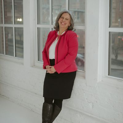 Smiling woman in red jacket and black skirt standing by windows.
