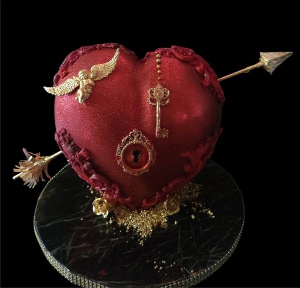 Red heart-shaped cake with golden key, lock, angel, and arrow on black base.