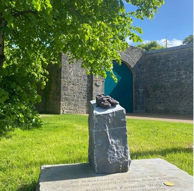 Memorial stone with shoes on top under green tree.